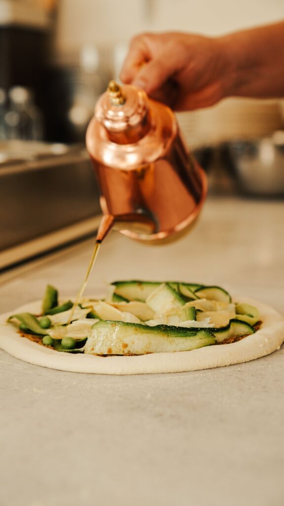 Aus einer kupernen Kanne wird Öl auf eine Pizza geträufelt.